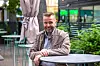 Mann sitter ved et rundt bord på en uteservering med planter og moderne bygninger bak.